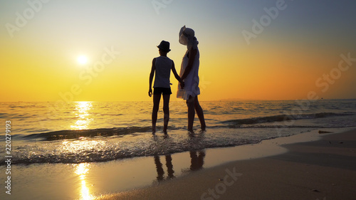 Mother with white dress and hat and son with hat standing barefoot on beach looking at the setting sun splashed by sea waves. Travel concept