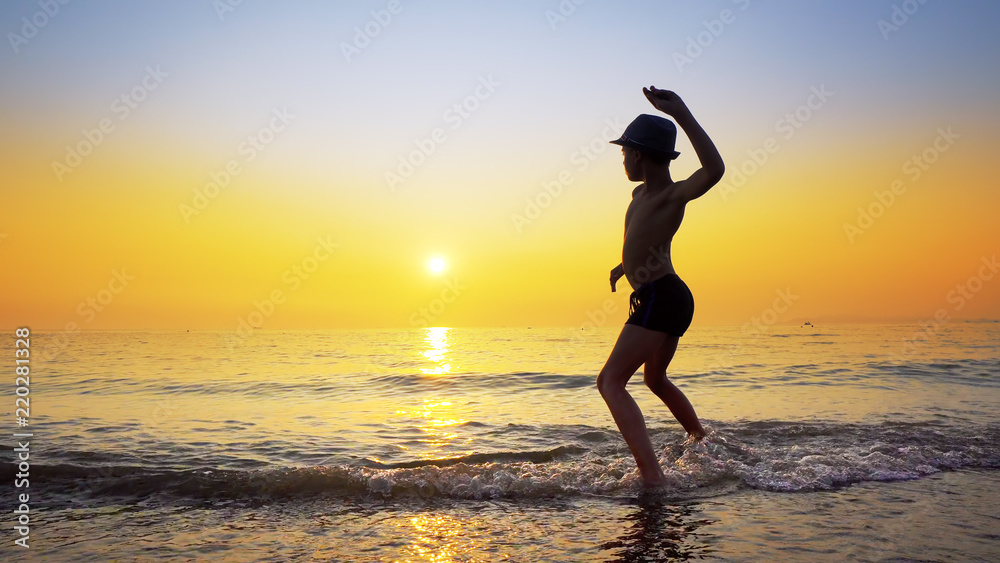 Silhouette of boy with hat throwing stones skipping on sea water surface. Summer vacation concept with vibrant orange sky