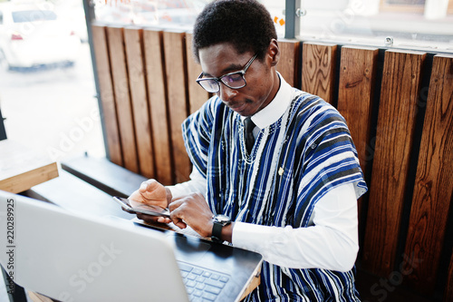 African man in traditional clothes and glasses sitting behind laptop at outdoor caffe with phone and looking at his watches.