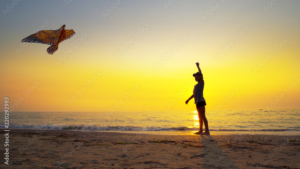 Silhouette of young boy with hat flying a kite on an empty beach backlit by summer sunset sun, cinematic footage