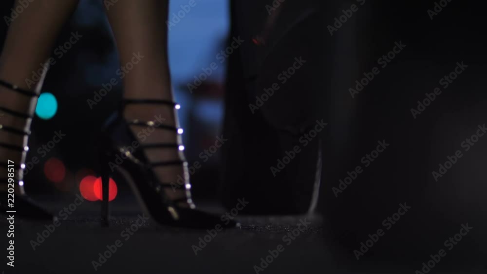 Close-up female legs in retro high heeled shoes aproaching automobile, opening car door and getting inside the car with elegance. City traffic lights bokeh on the background