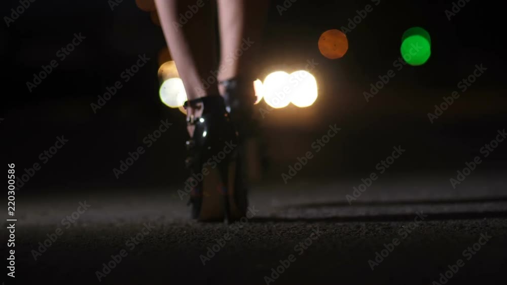 Close-up woman's feet and legs in black high heeled stiletto shoes walking at night on bokeh cityscape background. Female in stylish heels strolling in the evening on the street. Back view, steadicam