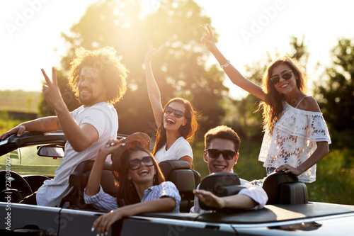 Beautiful stylish young girls and guys in sunglasses are sitting and laughing in a black cabriolet on a sunny day.
