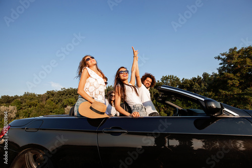 Dark-haired young women and curly dark-haired young man are standing in a black cabriolet on a summer day.