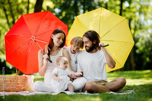 Kids and their parents sitting on the blanket under the big red and yellow umbrellas covering them from the sun.