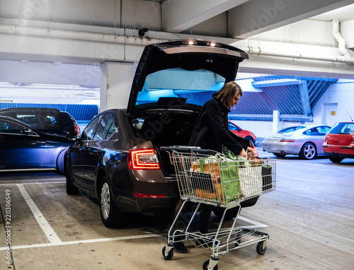 Side view of woman with shopping trolley near car putting bags with groceries into trunk on parking lot photo