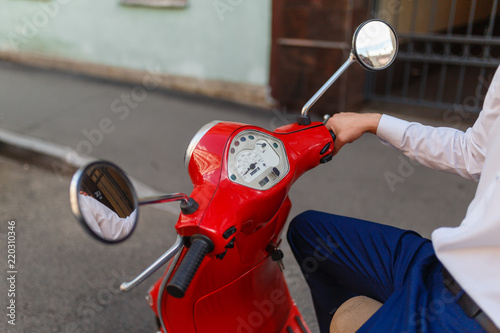 A man driving a red classic scooter rides on the asphalt in the city. Close - up of man's hands holding on to the red scooter's gas handles