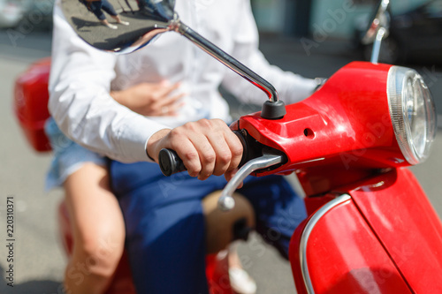 Man and woman on a classic scooter rides on the asphalt road. Girl hugging the man's waist sitting on the rear seat of the retro scooter. Closeup man hands holding on to the red scooter's gas handles