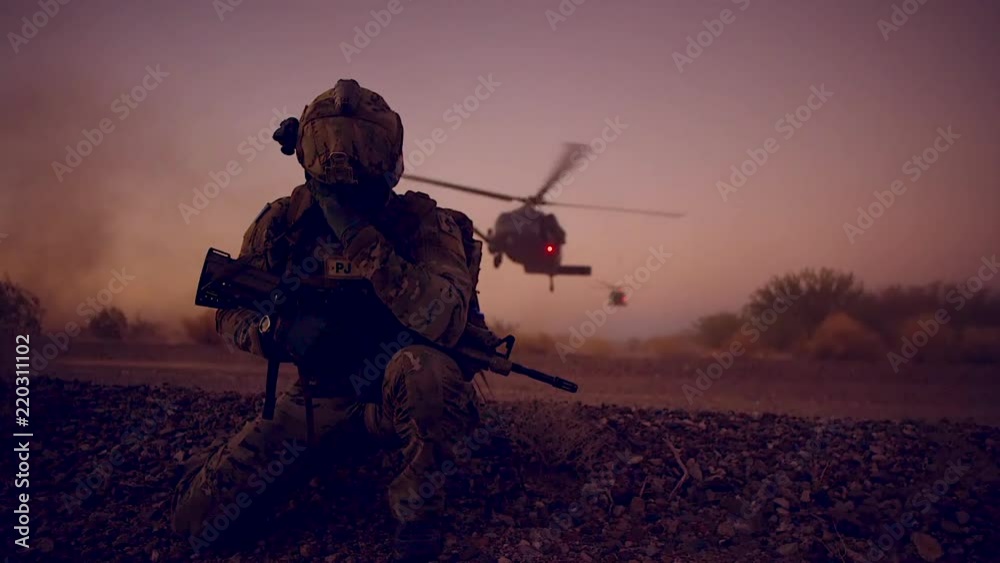 A soldier kneels as helicopters land on a mission behind him.