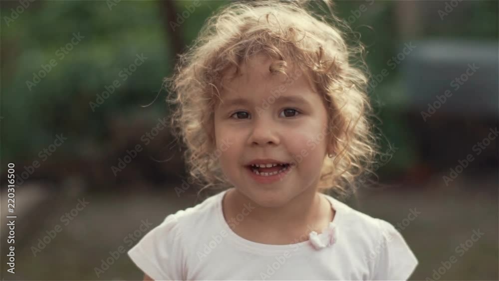 Portrait of the little pretty girl with long hair waving her hand in gront of the camera and smiling. Sunny green park on the background. Summer day. Slow motion Portrait of the little pretty girl with long hair waving her hand in gront of the camera and smiling. Sunny green park on the background. Summer day. Slow motion