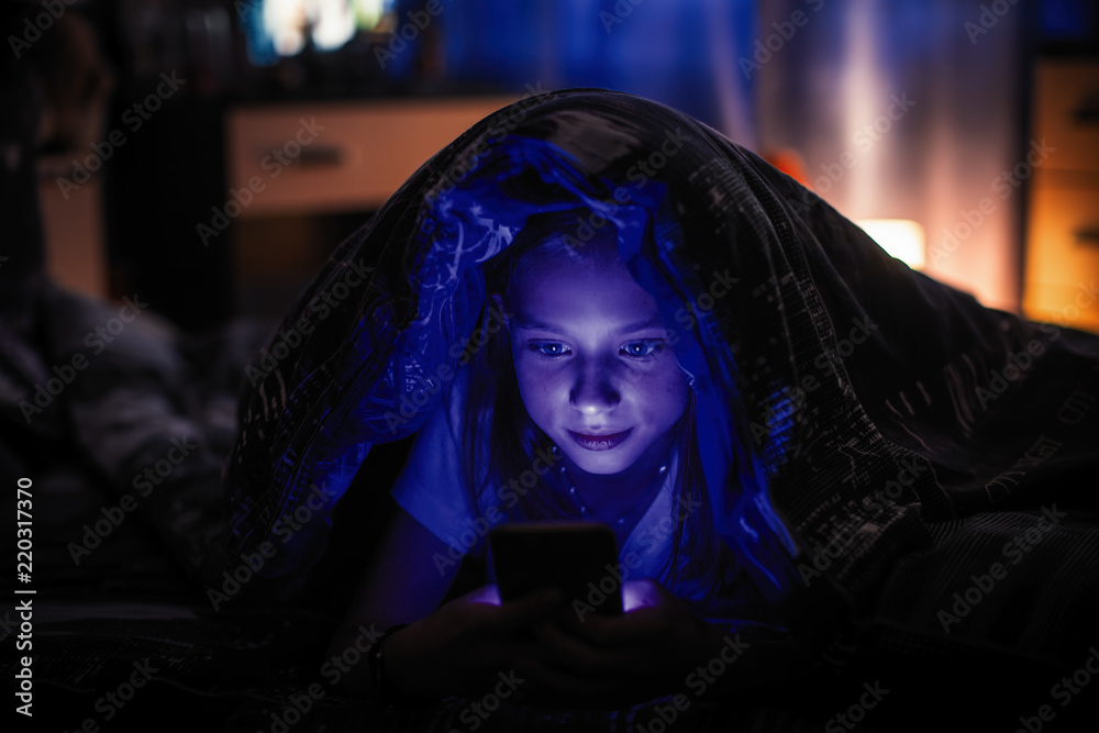 Teenage girl lying in the dark under a blanket looking at the smartphone. Teenage girl lying in the dark under a blanket looking at the smartphone.