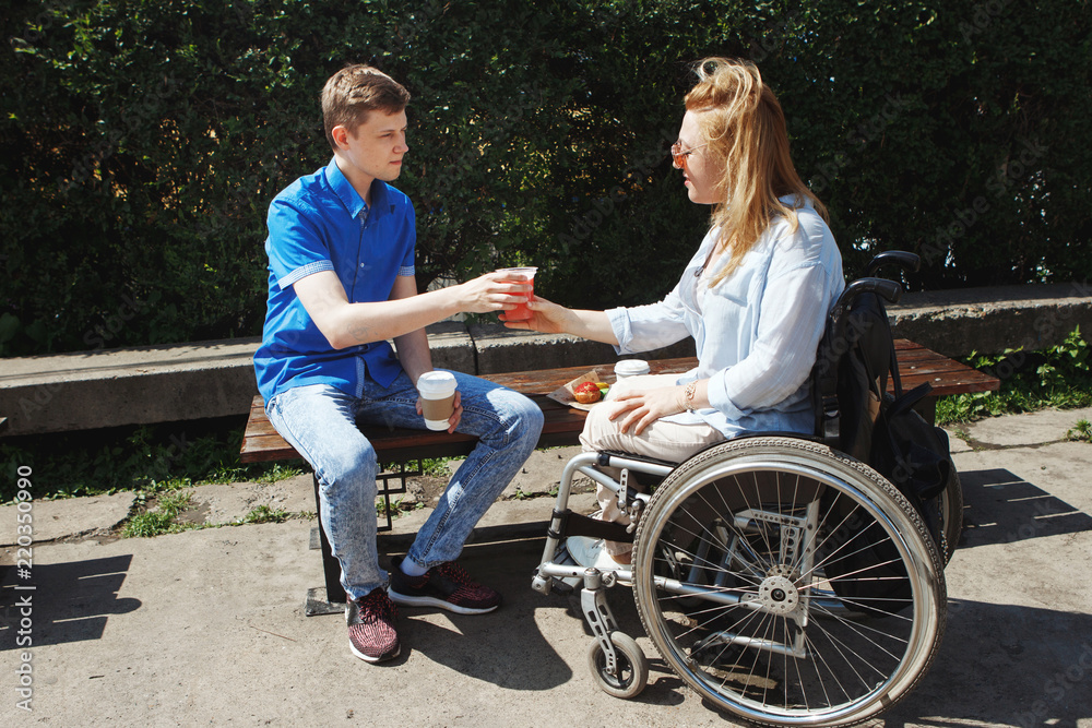 Woman in a wheelchair with her boyfriend drinking coffee and eating muffins in the city's summer park. Woman in a wheelchair with her boyfriend drinking coffee and eating muffins in the city's summer park.