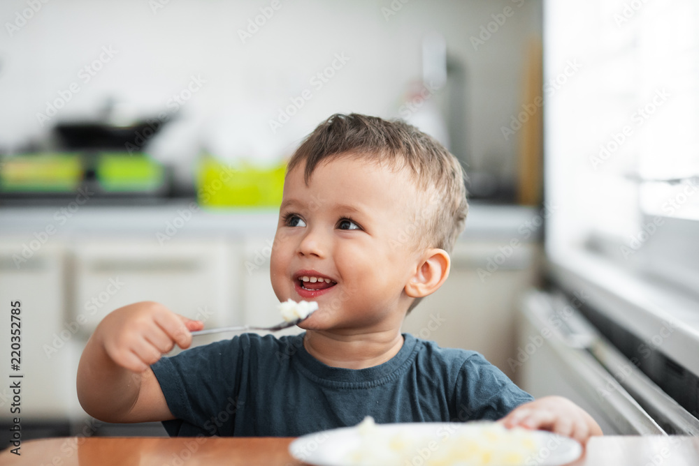 The little boy in the kitchen eagerly eating rice with a spoon independently The little boy in the kitchen eagerly eating rice with a spoon independently