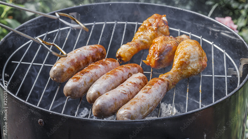.Grilled sausages and chicken legs on the grill. .Delicious.tasty juicy roast meat. Cooking food outdoor. process of cooking and rotate sausages with grilling tongs