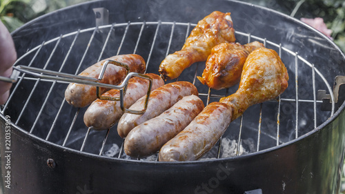 .Grilled sausages and chicken legs on the grill. .Delicious.tasty juicy roast meat. Cooking food outdoor. process of cooking and rotate sausages with grilling tongs