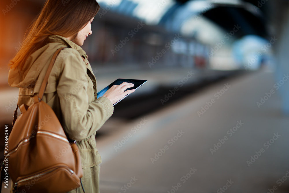 Fototapeta premium Young woman with backpack using tablet while standing on the railway station platform.