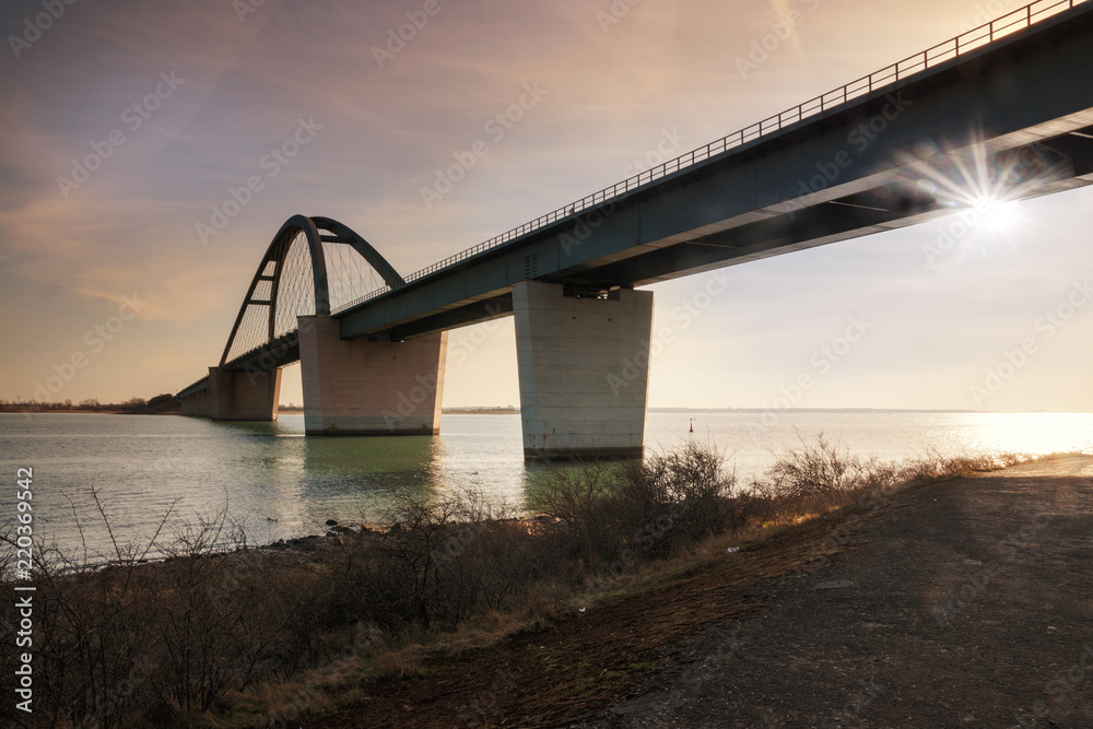 Fehmarn Sound Bridge in sunset (German: Fehmarnsundbrücke), suspension bridge with steel arches connecting the German mainland with the island in the Baltic Sea and belongs to the Vogelfluglinie Fehmarn Sound Bridge in sunset (German: Fehmarnsundbrücke), suspension bridge with steel arches connecting the German mainland with the island in the Baltic Sea and belongs to the Vogelfluglinie