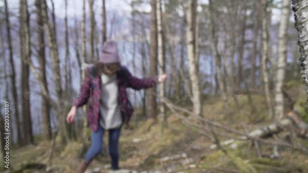 Woman with glasses, red cap, black and red jacket, grey sweater, blue backpack, blue jeans and brown boots hiking among trees with a fjord in the background_slomo Woman with glasses, red cap, black and red jacket, grey sweater, blue backpack, blue jeans and brown boots hiking among trees with a fjord in the background_slomo