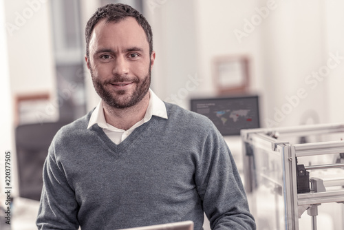 Feeling glad. Portrait of handsome bearded man smiling standing at the office with 3D printer on the background