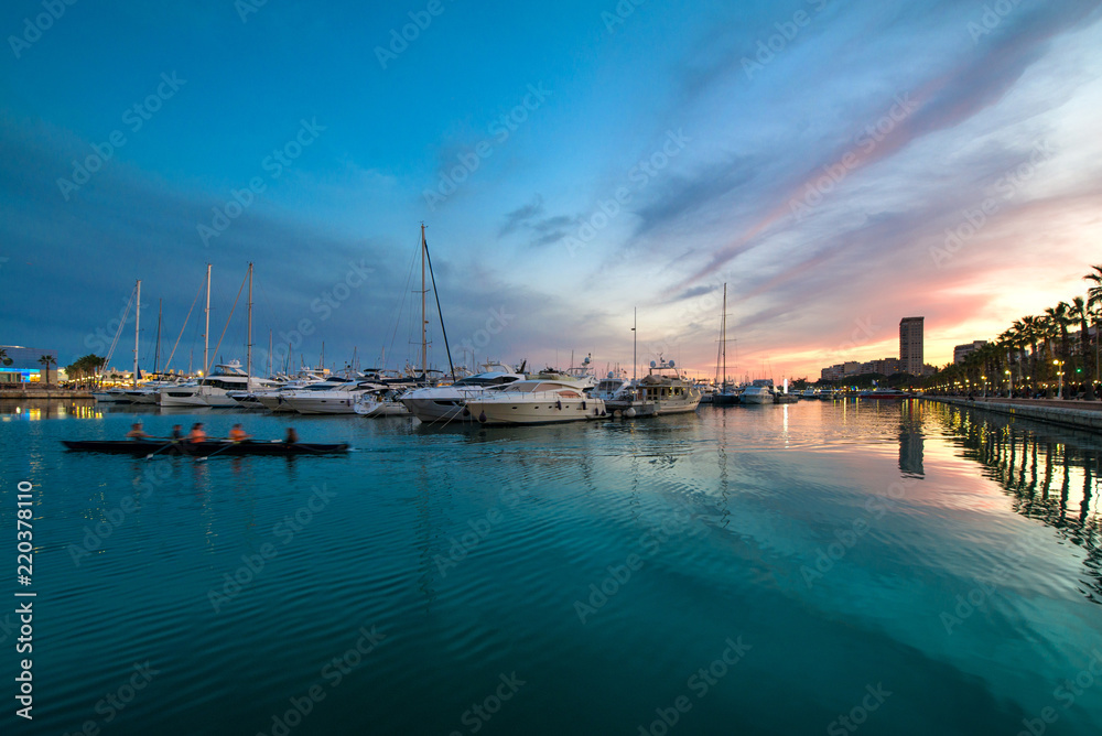 Obraz premium Beautiful port of Alicante, Spain at Mediterranean sea. Luxury yachts, ships, ferries and fishing boats sailing and standing in rows in harbor. Rich people traveling around the world. Sunset evening