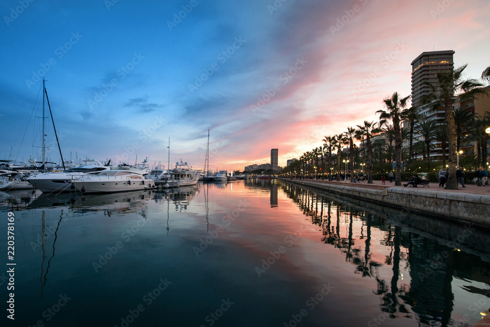 Beautiful port of Alicante, Spain at Mediterranean sea. Luxury yachts, ships, ferries and fishing boats sailing and standing in rows in harbor. Rich people traveling around the world. Sunset evening