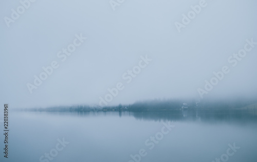 Cottages and villas seen through the dense fog covering water of lake. Shadows of the trees reflected in water. Sleepy misty landscape