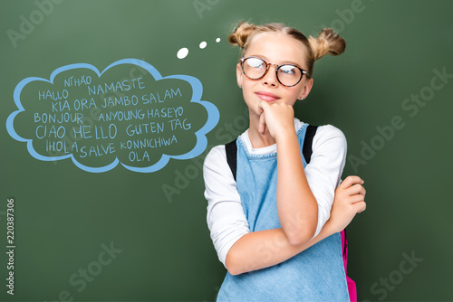 pensive schoolchild in glasses looking up near blackboard with words on different languages in speech bubble