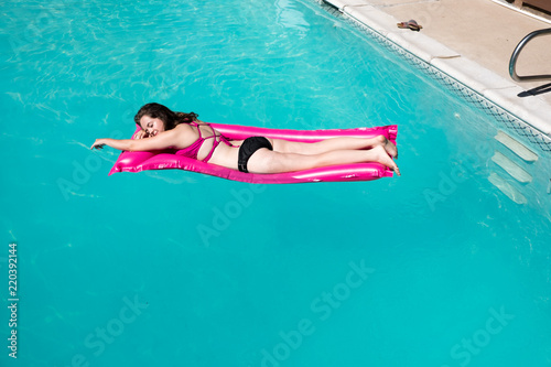White girl laying on stomach on pink raft. Enjoying lounging in an outdoor swimming pool, blue water and edge of pool. Caucasian girl in bikini on raft in pool arm extended sandals on edge of pool.
