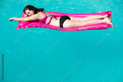 White girl laying on stomach on pink raft. Enjoying lounging in an outdoor swimming pool, blue water and edge of pool. Caucasian girl in bikini on raft in pool arm extended eyes closed. © Heather Shimmin