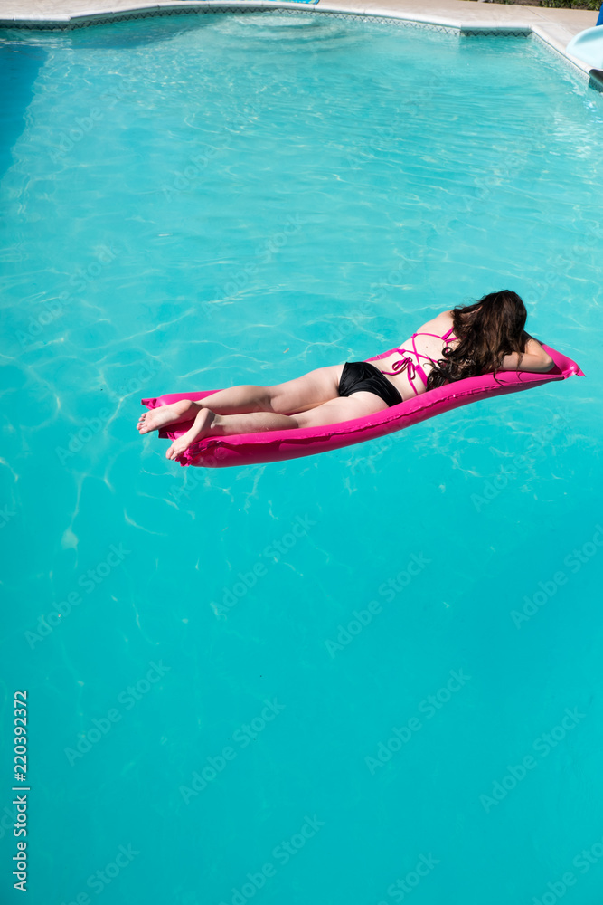 Looking down on a woman laying on her stomach on a pink raft wearing a bikini in a swimming pool. Yong woman on a raft in a swimming pool view from above.