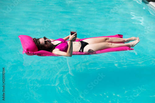 Pretty woman in a black and pink bikini and aviator sunglasses having a cocktail in an outdoor swimming pool on a bright pink raft. Woman floating in the swimming pool on raft in the summer sun.