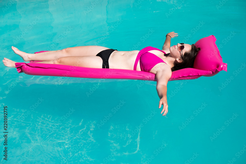 Pretty woman in a black and pink bikini  lounging in a swimming pool on a bright pink raft. Woman floating in the swimming pool on raft in the summer. Enjoying summer vacation.