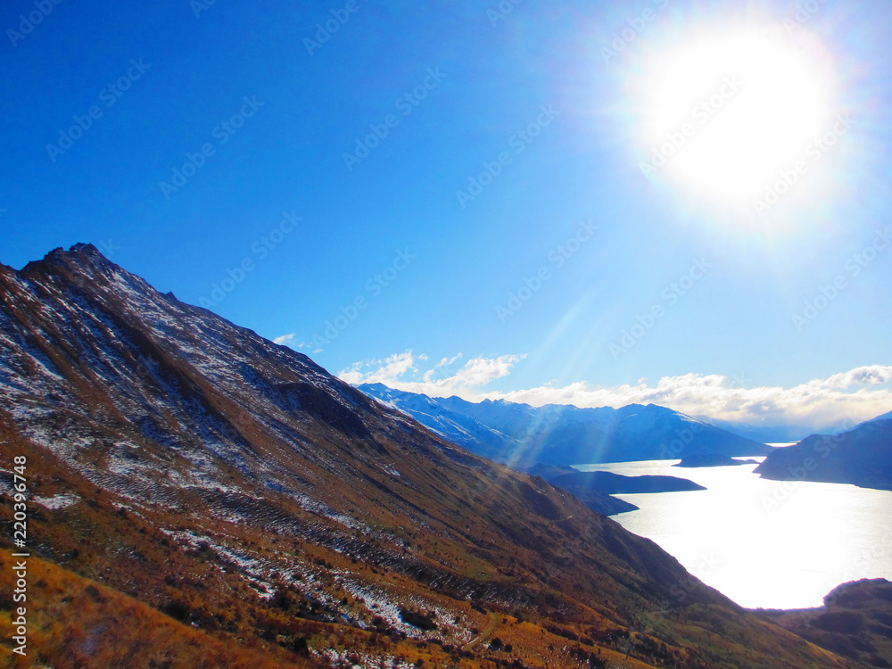 Image of Lake Wanaka, the mountain ranges and the sun in the sky. Taken from the side of Roys Peak in New Zealand on a clear winters day, with snow and grass in the foreground.