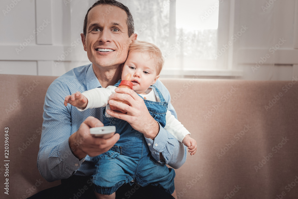 Interesting program. Cheerful positive man enjoying a TV show while holding his baby