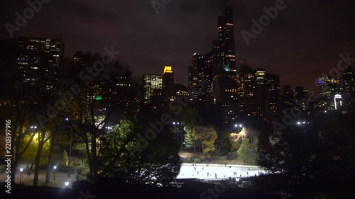 Beautiful view of people circling ice skating rink in Central Park at night under manhattan's skyline.