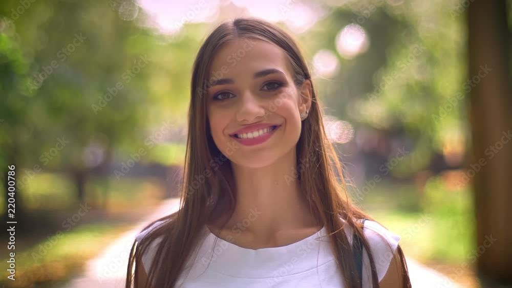 Nice lady is standing in park in daytime, watching at camera, smile, blurred backgrouns Nice lady is standing in park in daytime, watching at camera, smile, blurred backgrouns