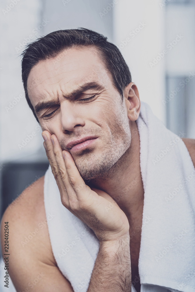 Pain. Exhausted personable man keeping his eyes closed and feeling sharp tooth pain while standing in the bathroom after having a shower Pain. Exhausted personable man keeping his eyes closed and feeling sharp tooth pain while standing in the bathroom after having a shower