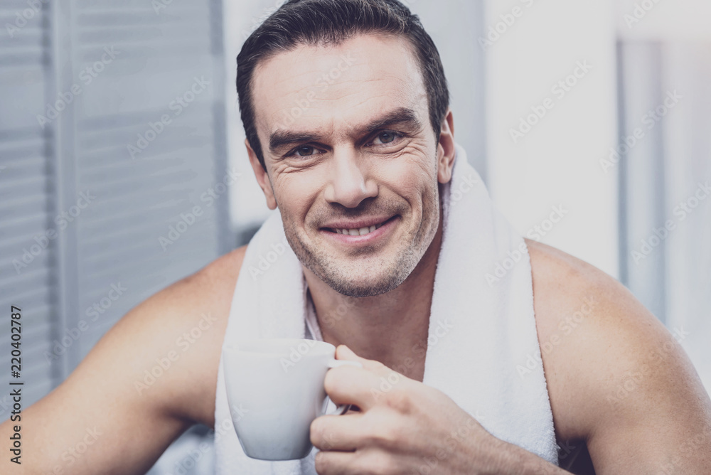 Pleasant morning. Optimistic delighted man with a towel around his neck holding a cup of hot coffee and having splendid mood while smiling at you Pleasant morning. Optimistic delighted man with a towel around his neck holding a cup of hot coffee and having splendid mood while smiling at you