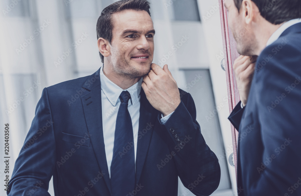 Titivation. Charismatic adorable entrepreneur feeling happy and looking at himself in the mirror while touching his chin with the hand