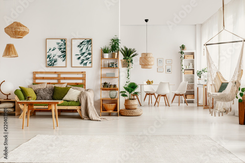 Posters above green sofa in white apartment interior with hammock and chairs and table. Real photo