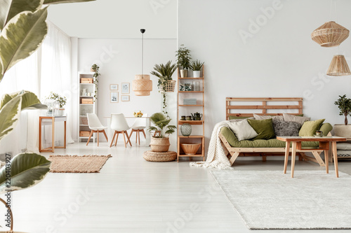 Wooden table in front of green couch in spacious living room interior with plants and lamps. Real photo