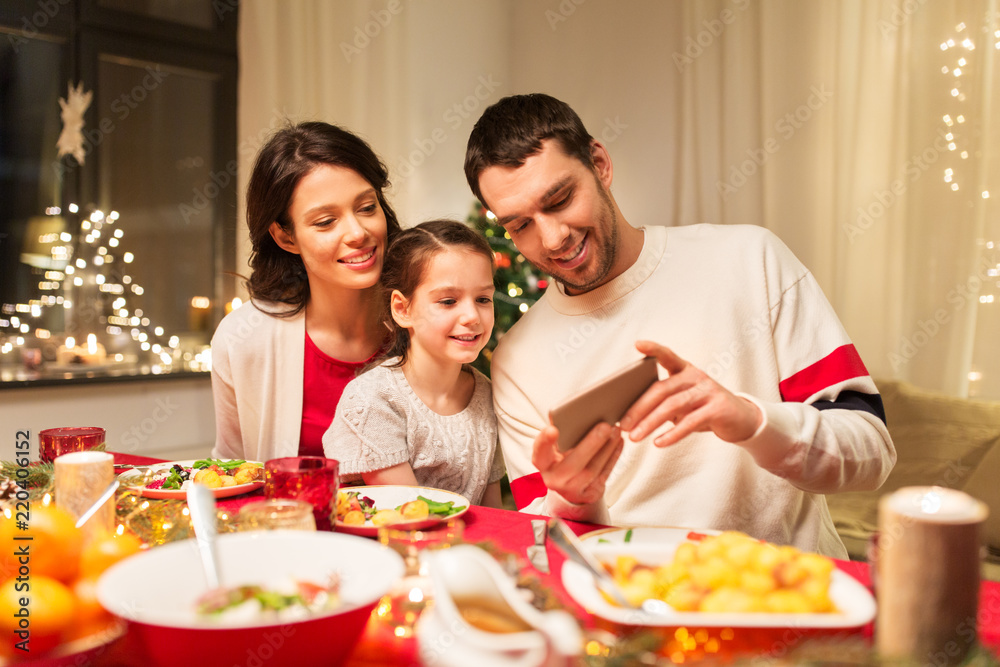holidays, family and technology concept - happy mother, father and little daughter with smartphone having christmas dinner at home