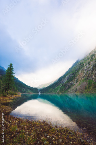 Amazing mountain lake in overcast weather. Mountains, cloudy sky and morning sunlight reflected in clear water. Coniferous trees near water. Stony bottom in transparent lake. Atmospheric landscape.
