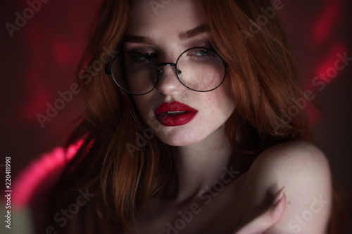 Redhead lady with freckless and long hair wearing eyeglasses looking aside over red lighting background.