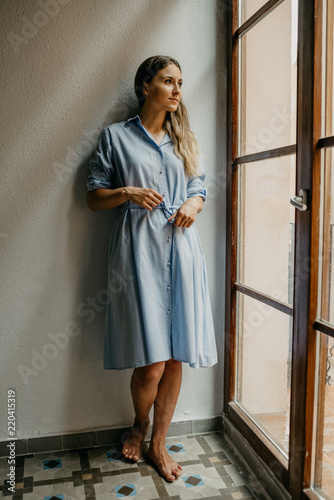 Pretty young women in the long blue dress posing on the floor of the flat with white walls looking out the window in Spain 