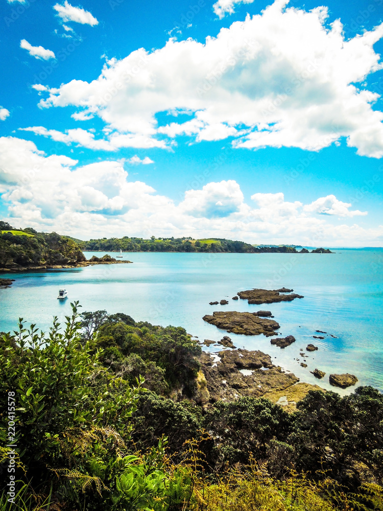 Hekerua Bay on Waiheke Island in New Zealand with sail boats on the water below and rocks and trees in the foreground. Taken on a bright Summer day.