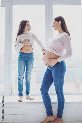 Child inside. Full length of pleased positive girl standing on window sill and touching her mothers belly while expressing delight on her face © Viacheslav Yakobchuk