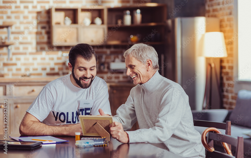 Look at this. Friendly senior satisfied man sitting by the table smiling and showing a photo to a volunteer. Look at this. Friendly senior satisfied man sitting by the table smiling and showing a photo to a volunteer.