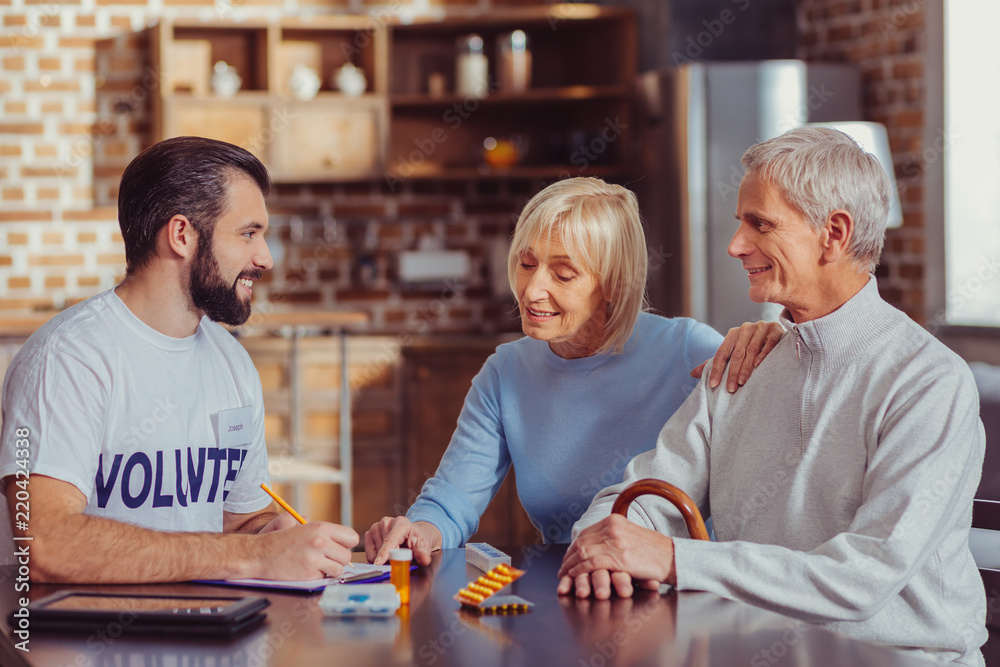 Fototapeta premium Explain please. Curious interested gray haired woman sitting by the table listening to a volunteer and pointing at notebook.