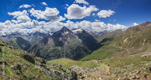 Wide panoramic view overlooking the mountain peaks, glaciers and rivers flowing down, forest on the sides of mountains, hiking trails, grass meadow on sunny summer day blue sky with clouds.